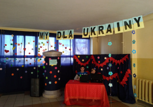 Szkolny korytarz, na którym dwóch chłopców siedzi przy stole, a nad nimi znajduje się napis: My dla Ukrainy.